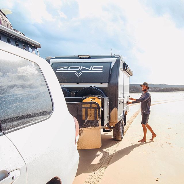 man standing besides a caravan hitched to a 4x4 on a beach