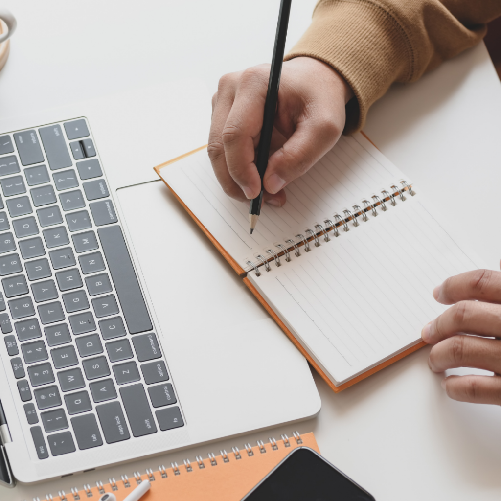 top view of a hand about to write in a note book next to a laptop