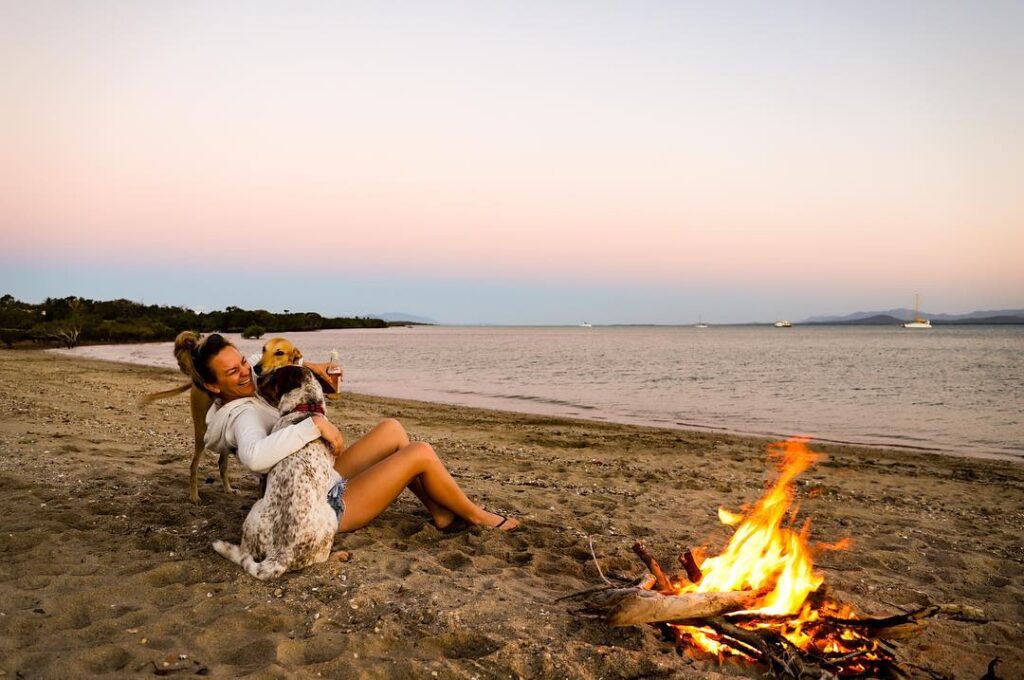 lady sitting on the beach hugging two dogs next to a camp fire