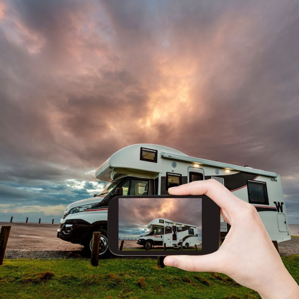 hand holding a phone in front of a motorhome with motohome on phone screen with a cloudy backround